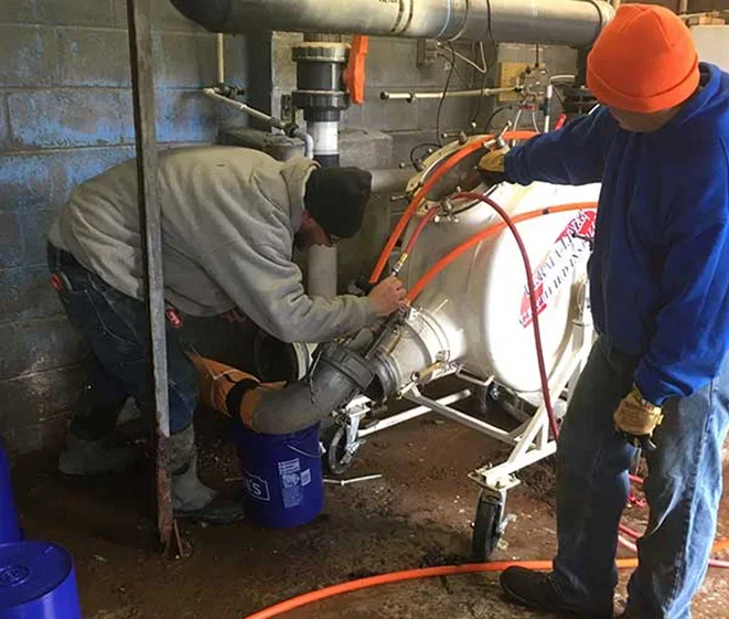 Two workers in a utility room are adjusting equipment connected to a large tank, while one holds an orange hose and the other operates a valve.