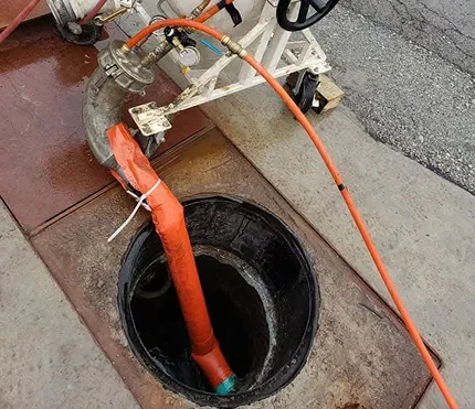 A close-up of a manhole with an orange hose inserted, connected to equipment for drainage or maintenance on a paved surface.