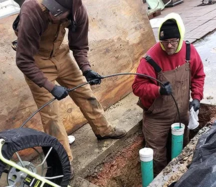 Two workers in brown overalls and gloves are operating equipment to inspect or clean sewage pipes in an open trench.